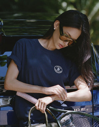 A woman wearing a navy blue t-shirt with a circular white logo on the left chest, styled outdoors with sunglasses and a tennis racket.