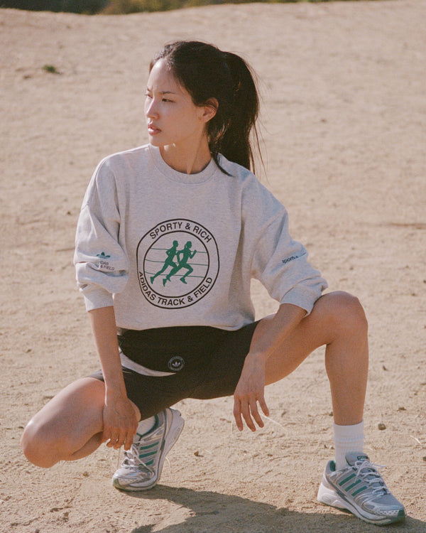 Person wearing a white sweatshirt with a logo, squatting on a sandy surface.