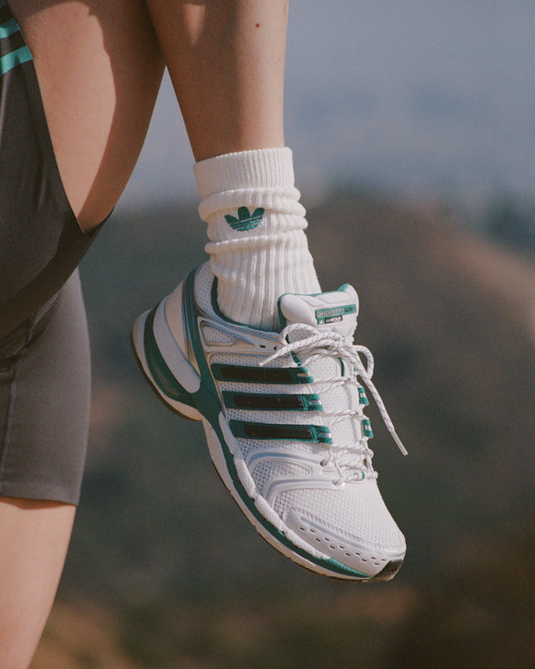 White Adidas sneaker with green stripes worn by a person on a blurred natural background