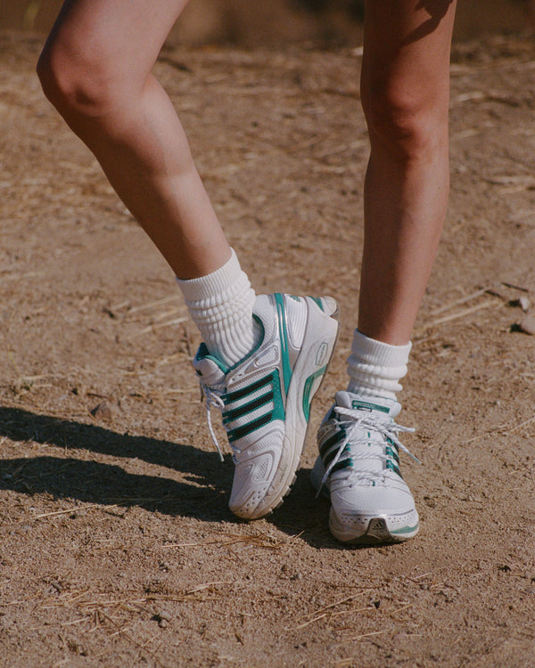 Close-up of two pairs of white sneakers with green accents on a sandy surface.