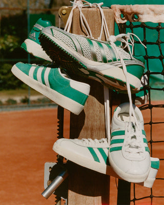 Three pairs of S&R Blanc - Court Green/White sneakers with premium suede uppers hang from a tennis net post on a clay court.