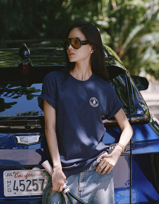 A woman wearing a navy blue crewneck t-shirt with a small circular white graphic on the left chest, paired with light-wash jeans, posing in front of a blue car outdoors.