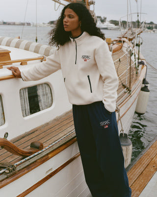 Woman wearing a white hoodie and navy pants with a logo, standing on a boat.