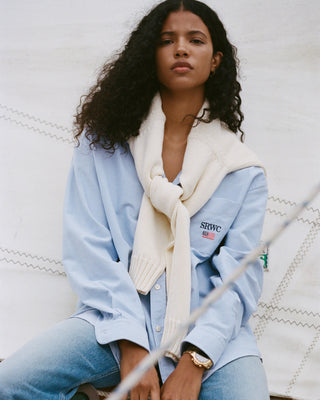Woman wearing a light blue shirt and jeans with a cream scarf, sitting against a white textured background.