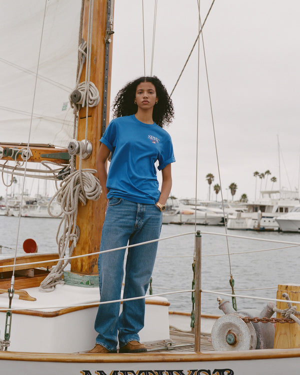 Person wearing a blue t-shirt and jeans standing on a boat with a marina in the background