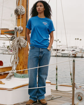 Woman wearing a blue t-shirt and jeans standing on a boat in a marina.