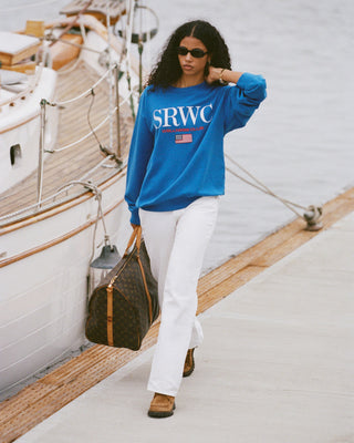 Woman in a blue sweatshirt with 'SRWC' text, white pants, and sunglasses standing on a dock with a boat in the background.