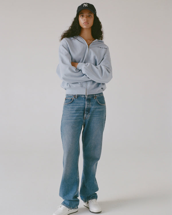 Person wearing a light gray hoodie, blue jeans, and a black cap on a plain background