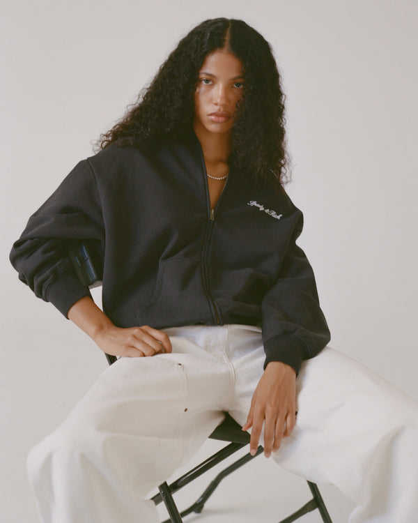 Person wearing a black jacket with a logo sitting on a chair against a white background