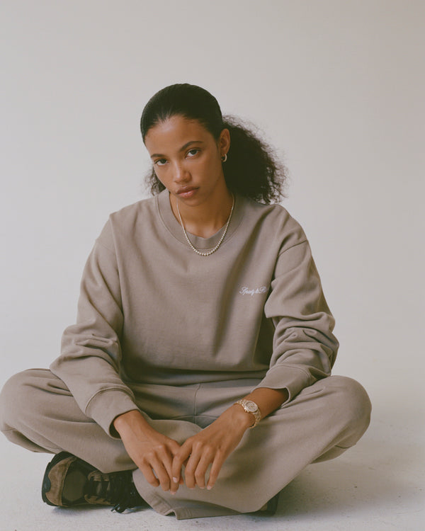 Person wearing a beige sweatshirt and pants sitting on the floor against a plain background