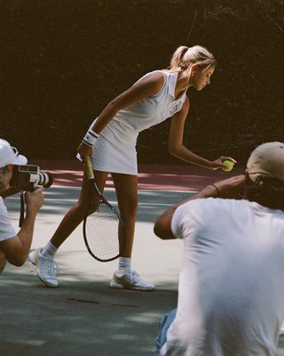 Woman in white tennis outfit preparing to serve a tennis ball on a court with photographers around.
