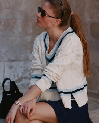 Woman wearing a white cardigan with black and blue trim, sitting against a stone wall.