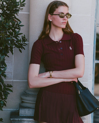 Woman in a maroon dress with a logo, sunglasses, and a black handbag standing against a light-colored wall.