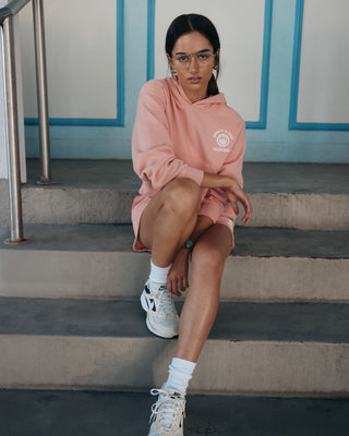 Woman wearing the California Emblem Soft Cropped Hoodie in Carnation/White and shorts sits on stairs, paired with glasses and white sneakers, looking at the camera.