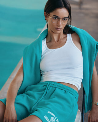 Woman in Varsity Crest Soft Gym Short - Spring Green/White, with a matching jacket, white tank top, and glasses, sitting by the poolside and looking at the camera.