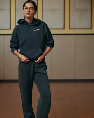 A woman stands in a gymnasium with one hand in her pocket, wearing glasses and the Serif Logo Soft Sweatpant in Faded Black/White with an elastic waistband.