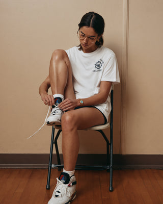A woman sitting on a chair ties her sneaker, wearing glasses and the California Emblem Soft T-Shirt in white, made in Portugal, paired with comfortable shorts.