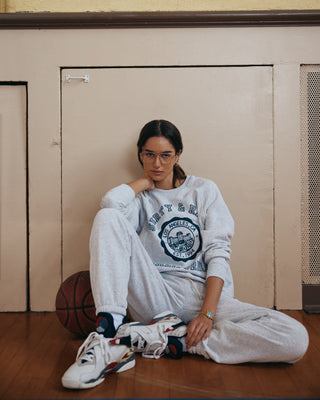 Woman in California Emblem Soft Sweatpant - Heather Gray/Navy sits indoors on the floor with a basketball, wearing glasses and sneakers.