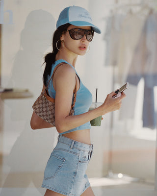 Woman in light blue top and denim shorts holding a drink and phone, wearing a blue cap.