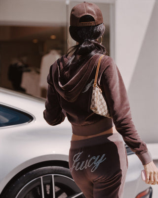 woman wearing a brown tracksuit with 'Juicy' text, standing next to a white car.