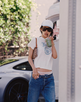 woman wearing a white t-shirt with a graphic design, blue jeans, and a white cap, standing next to a car.