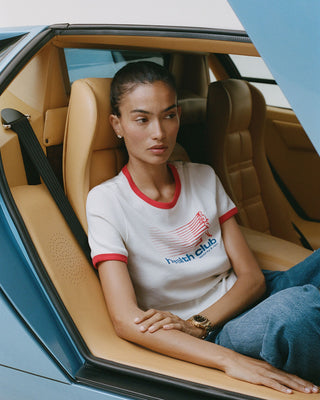 Woman sitting in a car wearing a white t-shirt with red trim and blue jeans.