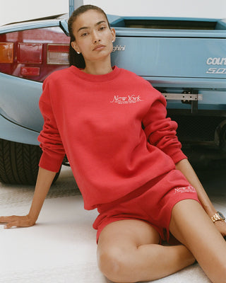Person wearing a red 'New York' sweatshirt and shorts sitting in front of a vintage car.