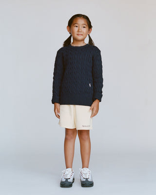 Child wearing a dark blue sweater and beige shorts on a plain background
