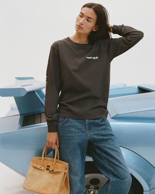 Person wearing a black sweatshirt with text, blue jeans, and holding a tan handbag in front of a blue car.