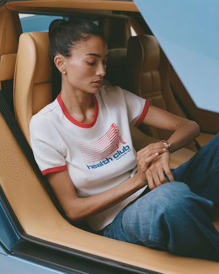 Woman sitting inside a car wearing a white t-shirt with red accents and blue jeans.