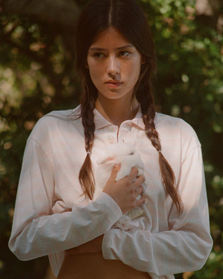 A young woman with dark braided hair wears a light pink long-sleeve collared shirt and holds a small white rabbit against her chest, set against a soft-focus green outdoor background.