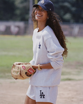 A woman outdoors on a baseball field wearing a light gray Los Angeles Dodgers sweatshirt and matching shorts, holding a tan baseball glove, with a navy cap featuring an LA logo.