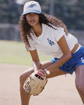 A woman on a baseball field wearing a white Los Angeles Dodgers t-shirt from the back, featuring the team's script logo and 'LA' logo in blue and red, paired with blue athletic shorts and a white cap.