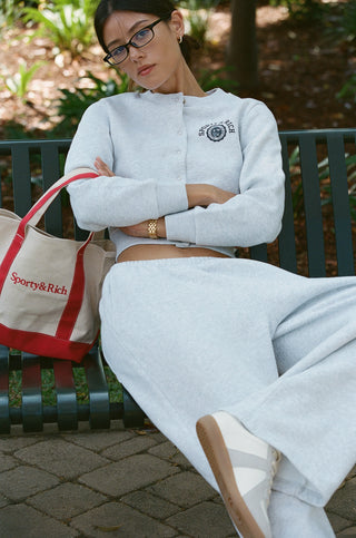 Woman in glasses wearing the Essia SR Emblem Fleece Cardigan in Heather Gray loungewear set and red flip-flops, standing against a white wall.