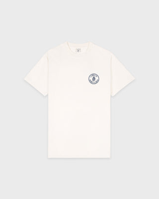 Ecru Cadillac x Sporty & Rich Atlas Tee with a small circular blue and black logo on the left chest, laid flat on a white background. Made from premium USA grown cotton, this tee offers timeless style and exceptional comfort.