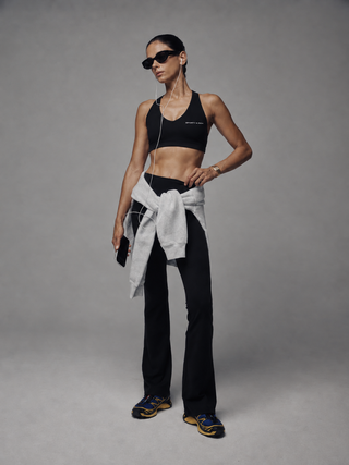 A black sports bra with a crossover V-neckline and double-strap back design, featuring the text 'SPORTY & RICH' printed in white on the lower left chest, displayed on a clean white background.