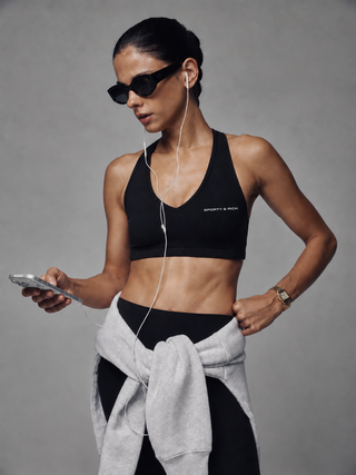 A black sports bra with a crossover V-neckline and double-strap back design, featuring the text 'SPORTY & RICH' printed in white on the lower left chest, displayed on a clean white background.