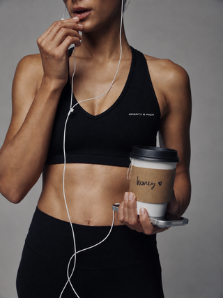 A black sports bra with a crossover V-neckline and double-strap back design, featuring the text 'SPORTY & RICH' printed in white on the lower left chest, displayed on a clean white background.