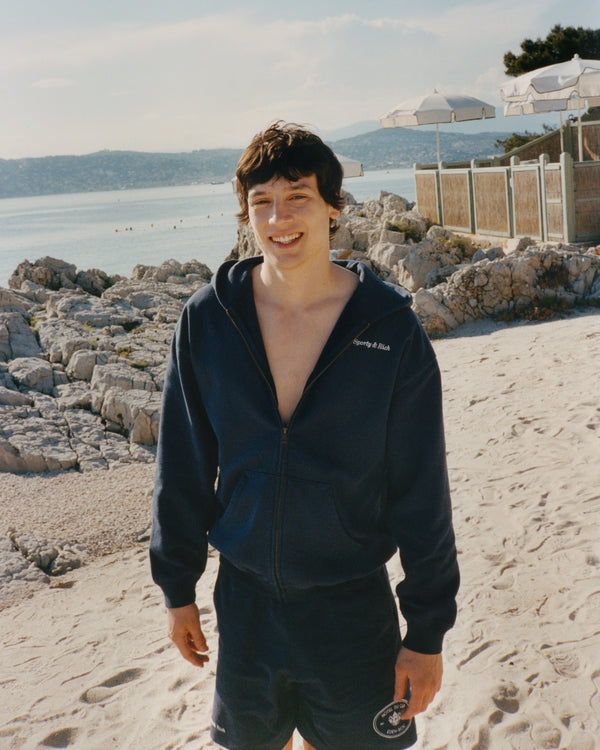 A smiling young person wears the Eden Crest Gym Short - Navy/White, standing on a sandy beach with rocks and the sea in the background.
