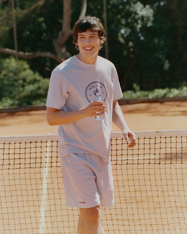 A smiling person in light attire, wearing Eden Crest Gym Short - Ciel/Navy with an elastic waistband, stands on a clay tennis court holding a drink in front of the net.