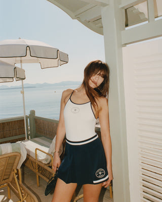 Woman wearing the Eden Crest Embroidered Mila Skirt in Navy/White and a white tank top poses on a sunny patio by the sea, with an umbrella and chairs nearby.