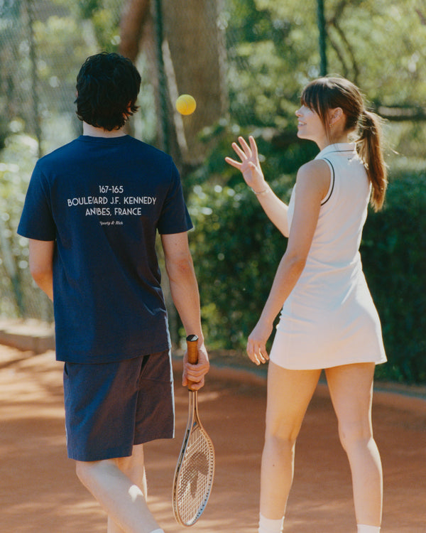 A woman in a Navy/White Kennedy Crest T-Shirt, made in Portugal, tosses a ball and smiles at a man on a clay tennis court.