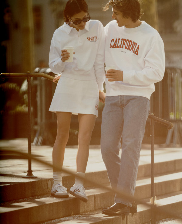 Two people stand on steps, smiling and holding coffee, both in casual white sweatshirts. One wears the Golf Logo Kelly Skirt - White/Bright Red, featuring an elastic waist and high-quality fabric.