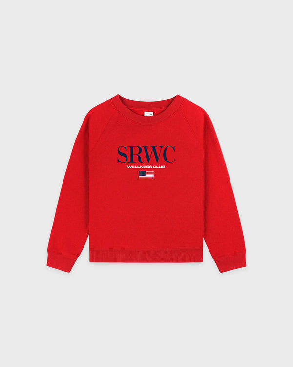 Nautical Kids Crewneck - Ruby: This red crewneck sweatshirt features "SRWC WELLNESS CLUB" and a small red-and-white striped graphic on the chest. Crafted from a soft cotton-polyester blend for comfort and durability, it offers an oversized fit and is made in Portugal—a cozy essential for kids.
