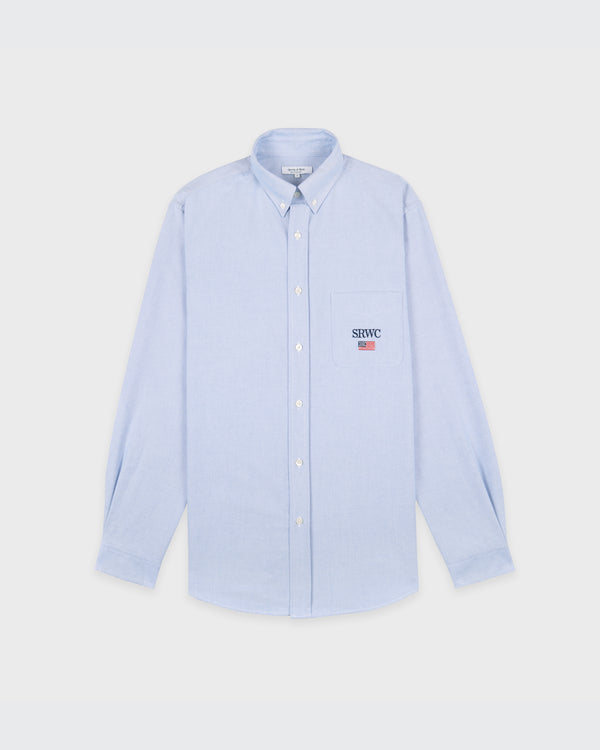 Nautical Oversized Oxford Shirt Men - Blue: An oversized, light blue Oxford Cotton shirt featuring a front pocket with an embroidered logo above it, elegant mother of pearl buttons, and presented laid flat on a white background.