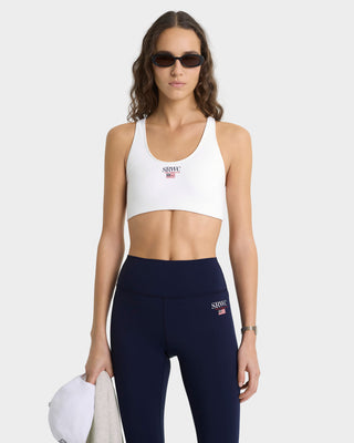 Woman wearing a white sports bra and navy leggings with a brand logo on a white background