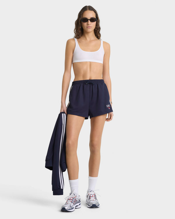 Woman wearing a white sports bra and navy shorts with a jacket on a white background
