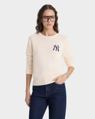Woman wearing a cream sweater with a navy logo on the chest and dark jeans on a white background