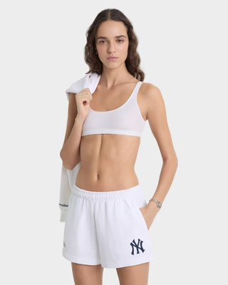 Woman wearing a white sports bra and shorts with a logo, holding a towel on a light gray background