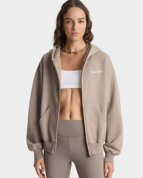 Woman wearing a beige hoodie and leggings with a white sports bra on a plain background
